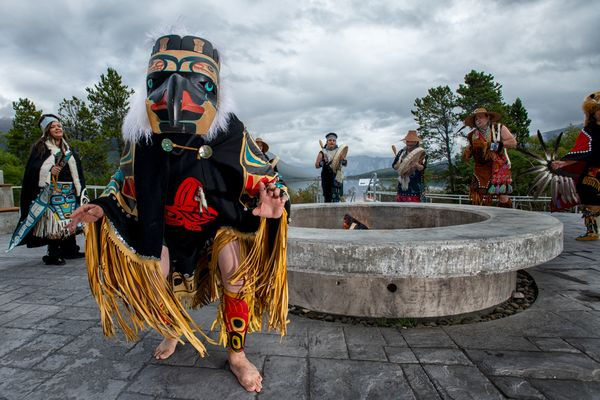 Person in colorful traditional attire and mask dances energetically. Others in similar outfits dance near a stone structure, set against a cloudy sky.