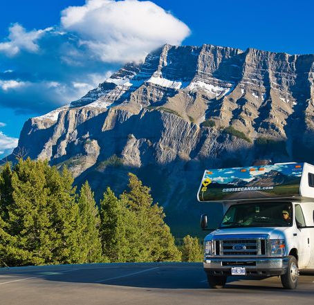 An RV parked on a road with majestic mountains in the background under a blue sky, surrounded by green trees.
