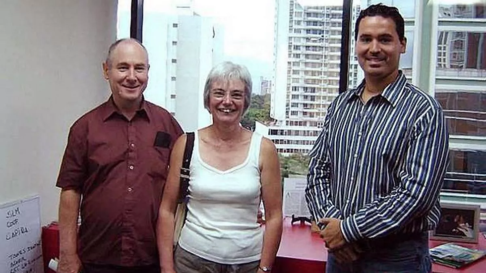 Three people smiling indoors, with tall buildings visible through the window behind them. A red desk with papers and books is in the foreground. John & Anne Darwin and real estate agent in Panama.