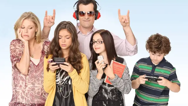 A group of five people stand together. Three are using devices, one holds a book, and one, wearing headphones, gestures playfully. Casual mood. Modern Family family.