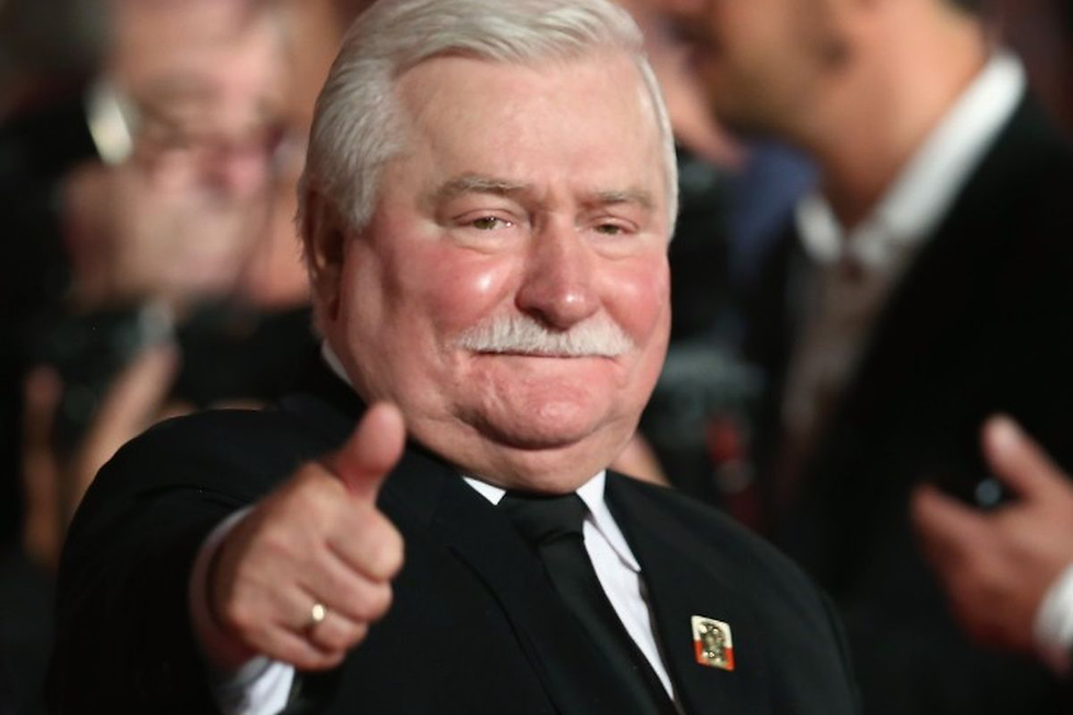Lech Walesa in a suit giving a thumbs-up, smiling confidently in a crowded setting. Close-up view with a soft-focus background.