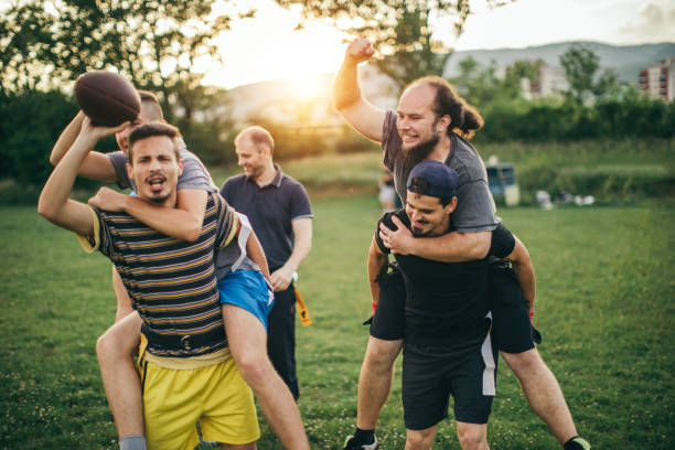 Men playing touch football in a green field at sunset. One man carries another, laughing and energetic. Trees and buildings in the background.