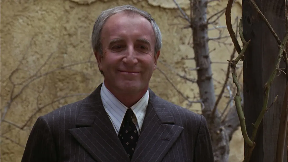 Smiling man in a pinstripe suit stands outdoors against a textured beige wall. Bare tree branches visible; calm and content mood. Peter Sellers as Chance the Gardener in Being There.