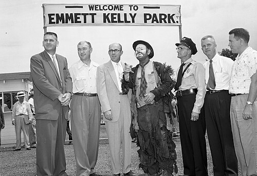 Group of men and a clown pose under a WELCOME TO EMMETT KELLY PARK banner at an outdoor ceremony.