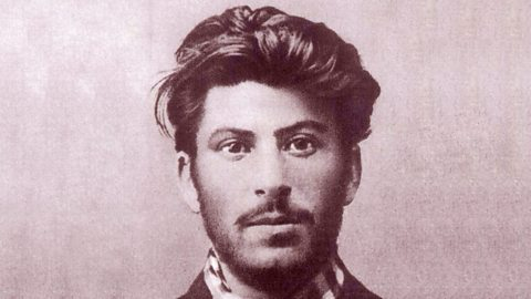 A sepia-toned portrait of a young Joseph Stalin with tousled hair, wearing a patterned scarf. The background is plain, creating a calm mood.