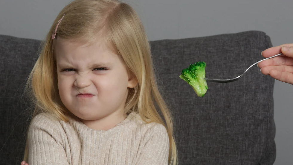Young girl grimaces as a hand offers her a forkful of broccoli on a gray couch, showing dislike or refusal