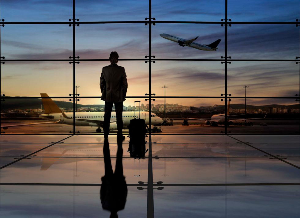 A person stands in an airport looking at planes on the runway. One plane takes off. Sunset sky and reflections on the floor create a serene mood.