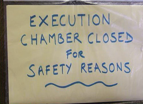 Sign reading "EXECUTION CHAMBER CLOSED FOR SAFETY REASONS" in blue text on yellow background, conveying urgency and caution.