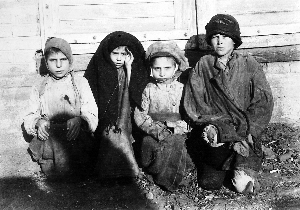 Four Russian children in worn clothing sit on the ground against a wooden wall. Their expressions are serious, and the setting appears rustic and bleak.