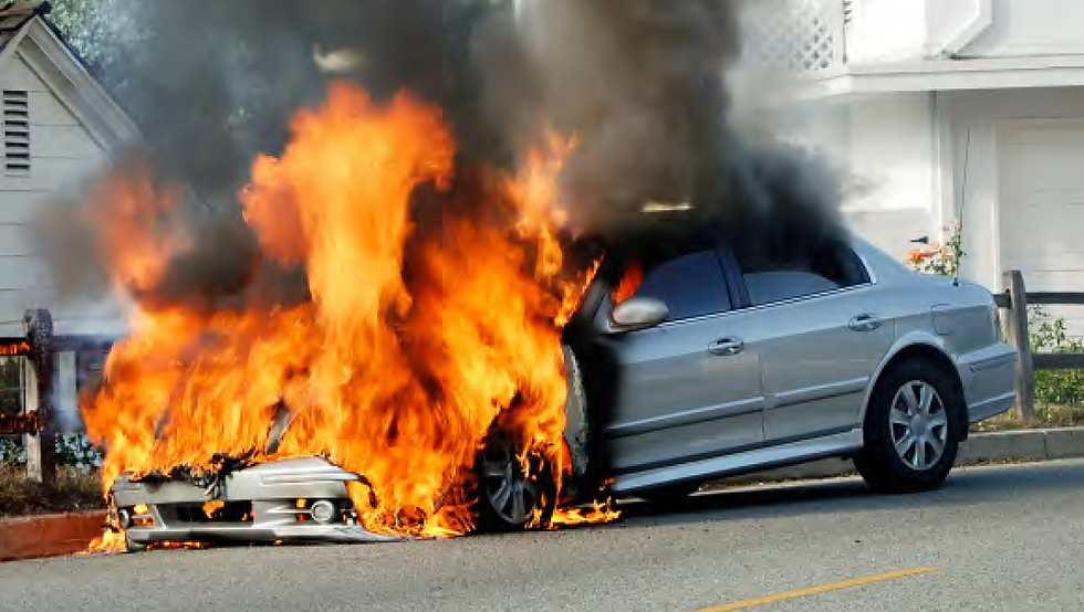Silver car engulfed in bright orange flames and black smoke, parked by a white house. Fiery scene, daytime setting.
