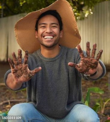 Smiling person outdoors wearing a wide-brimmed hat and sweater, showing muddy hands, with a garden and fence in the background.