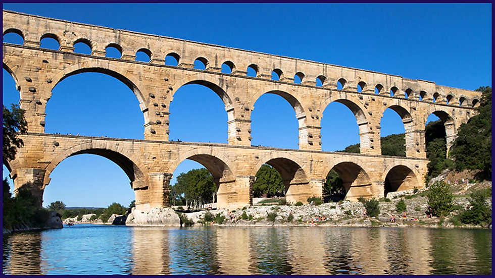 Ancient stone aqueduct with multiple arches spans a river under a clear blue sky. Trees and people are visible along the riverbank.