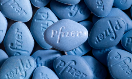 Close-up of numerous blue tablets with "Pfizer" embossed. Tablets form a layered pattern, set on a dark background. Viagra.