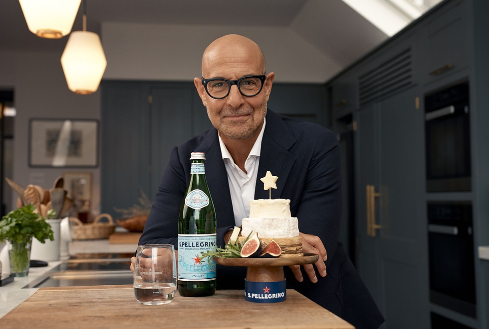 Man in glasses, Stanley Tucci, with a friendly expression holding a wooden tray with a small cake topped by a star, figs, and a bottle of San Pellegrino. Kitchen setting.