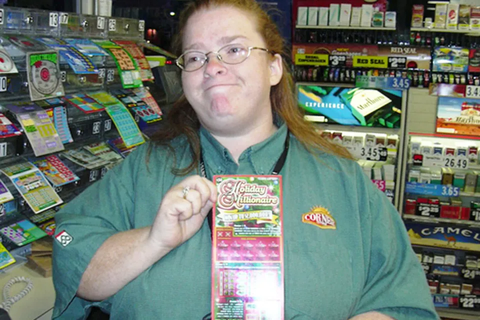 Store employee in a green shirt holding a "Holiday Millionaire" scratch-off card, surrounded by lottery tickets and tobacco products on shelves. Joan Ginther.
