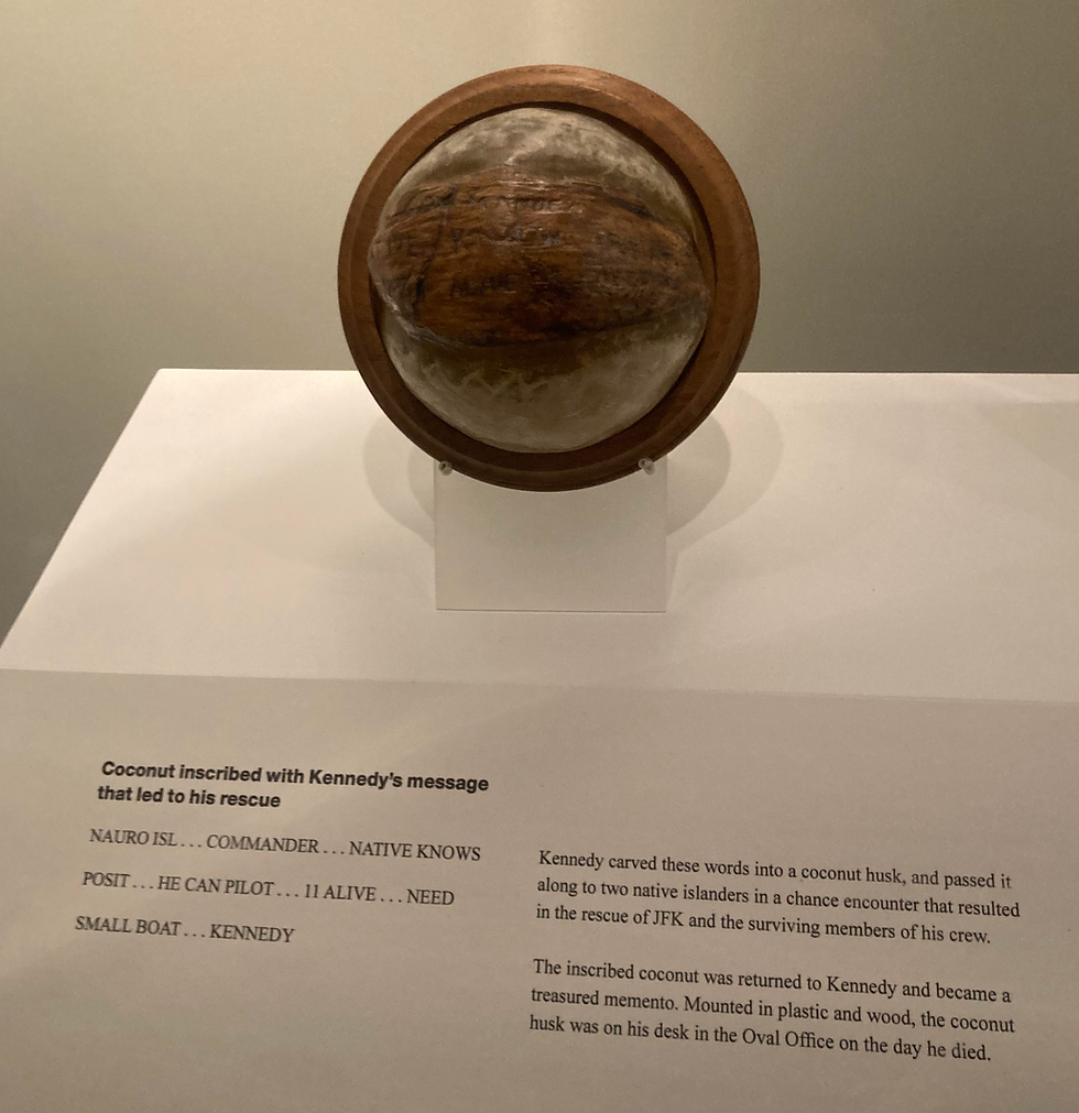Coconut inscribed with rescue message in display, surrounded by text describing its significance for JFK. Mounted on wood and plastic. Neutral setting.
