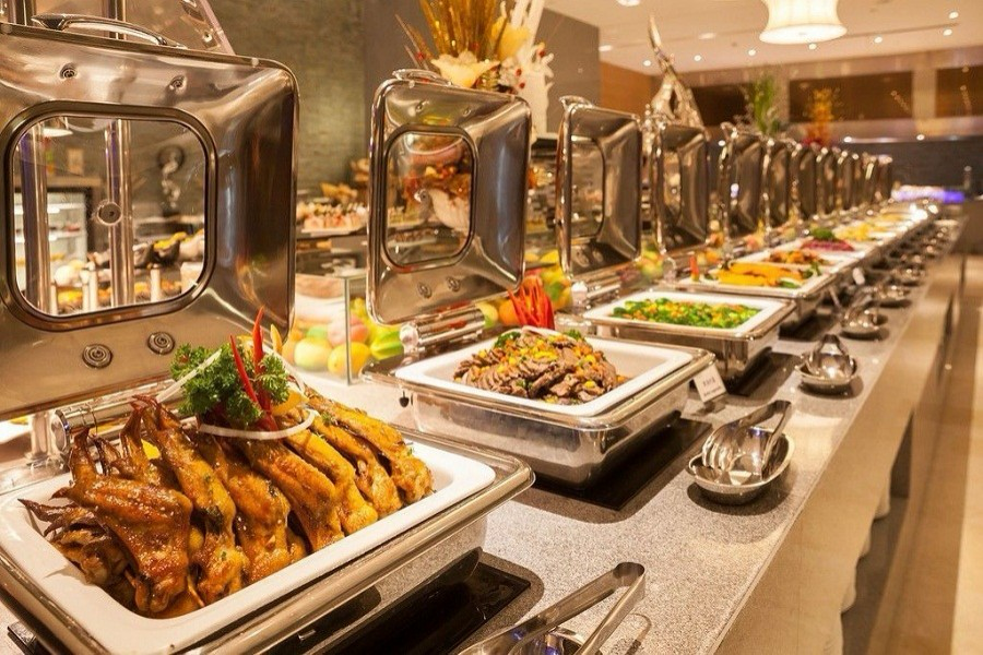 Buffet table with trays of gourmet dishes, including roasted poultry and vegetables. Elegant restaurant setting with stainless steel accents.