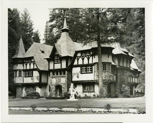 Black-and-white photo of a Tudor-style mansion in the woods, with a statue on the front lawn. Wyntoon.