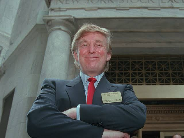 Donald Trump smiles with arms crossed outside the New York Stock Exchange, wearing a suit, red tie, and name tag.