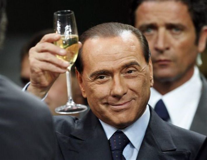 Silvio Berlusconi toasting with a champagne glass, smiling in a formal suit. Another man in the background appears serious. Indoor setting, dim lighting.
