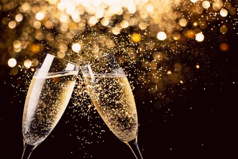 Two champagne glasses clink, surrounded by golden bokeh lights on a dark background, creating a festive and celebratory mood.