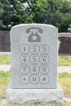 A tombstone shaped like a phone keypad with numbers and letters. It's set in a grassy cemetery; mood is humorous or ironic.