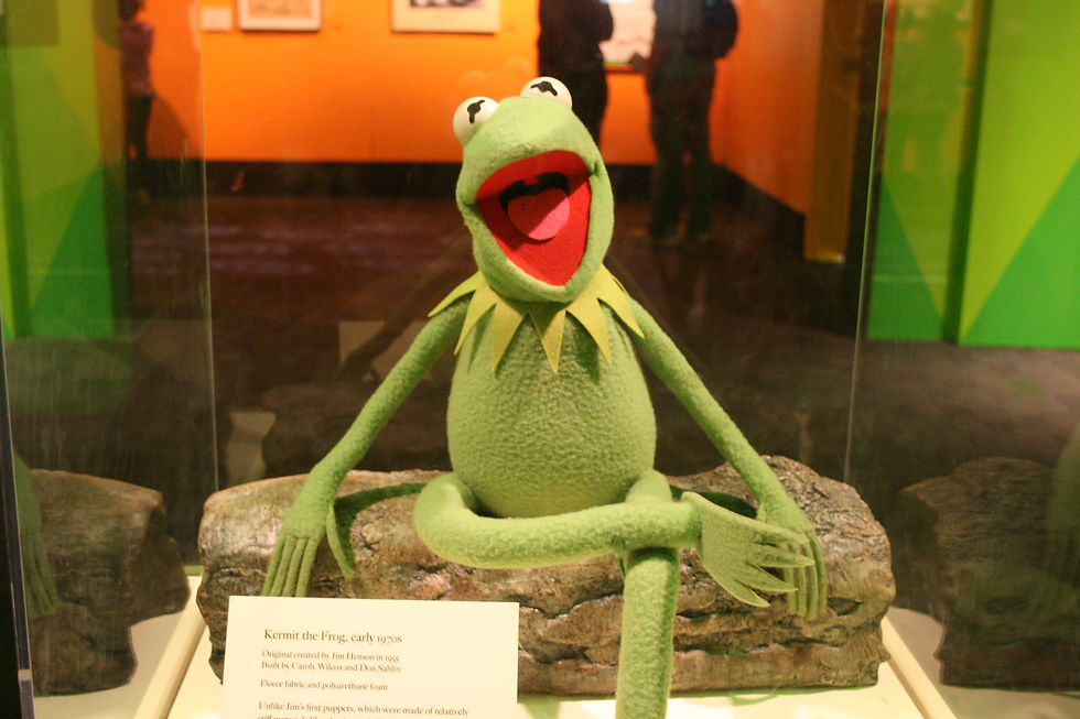 Kermit the Frog puppet displayed with a cheerful expression in a glass case, set against an orange background with informational text below.
