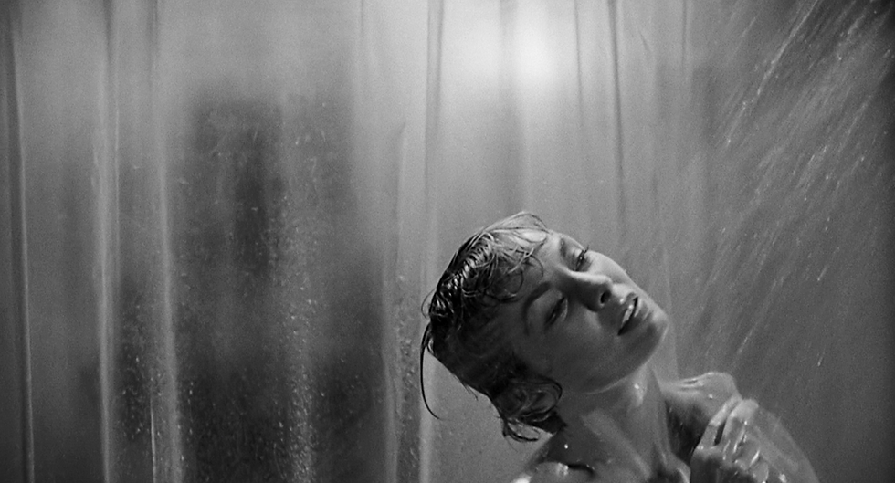 A person is taking a shower, head tilted back under the water. A shadowy figure is visible through the shower curtain, creating tension.