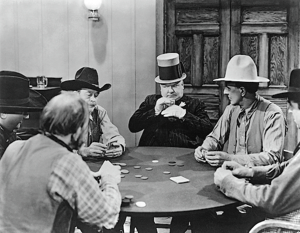 Black-and-white scene of cowboys playing poker around a table, one man smoking, in a rustic room with wooden doors. W.C. Fields