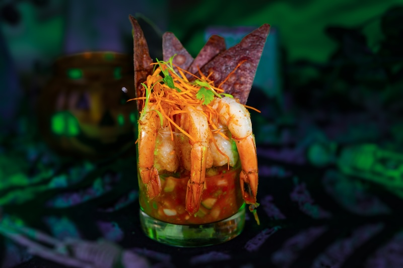 Shrimp cocktail in a glass, garnished with shredded carrots and cilantro, surrounded by purple chips. Background has green and purple hues.