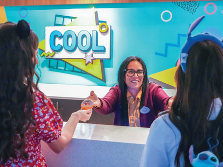 Smiling employee at a colorful desk gives a button to two women, one in a red dress, the other with ears headband. Sign reads "COOL".