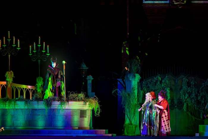 Characters in colorful costumes perform on a dimly lit stage with gothic elements. One holds a staff; others look surprised. Dramatic atmosphere.