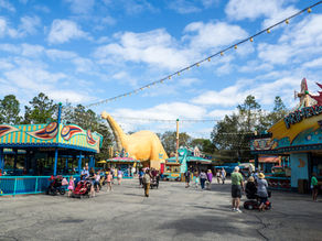 People stroll in a colorful amusement park with a large dinosaur statue, vibrant stalls, and string lights under a partly cloudy sky.