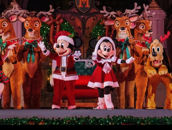 Mickey and Minnie in festive outfits dance with reindeer and Pluto on a decorated stage. Christmas lights and colors create a joyful mood.