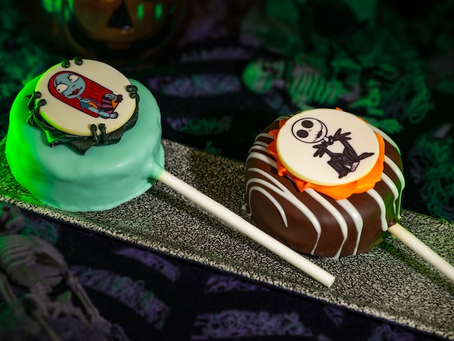 Brownie and yellow cake pops dipped in chocolate with a candied picture of Jack Skellington and Sally Skellington on them. These are sitting on a small serving platter with a spooky themed background.