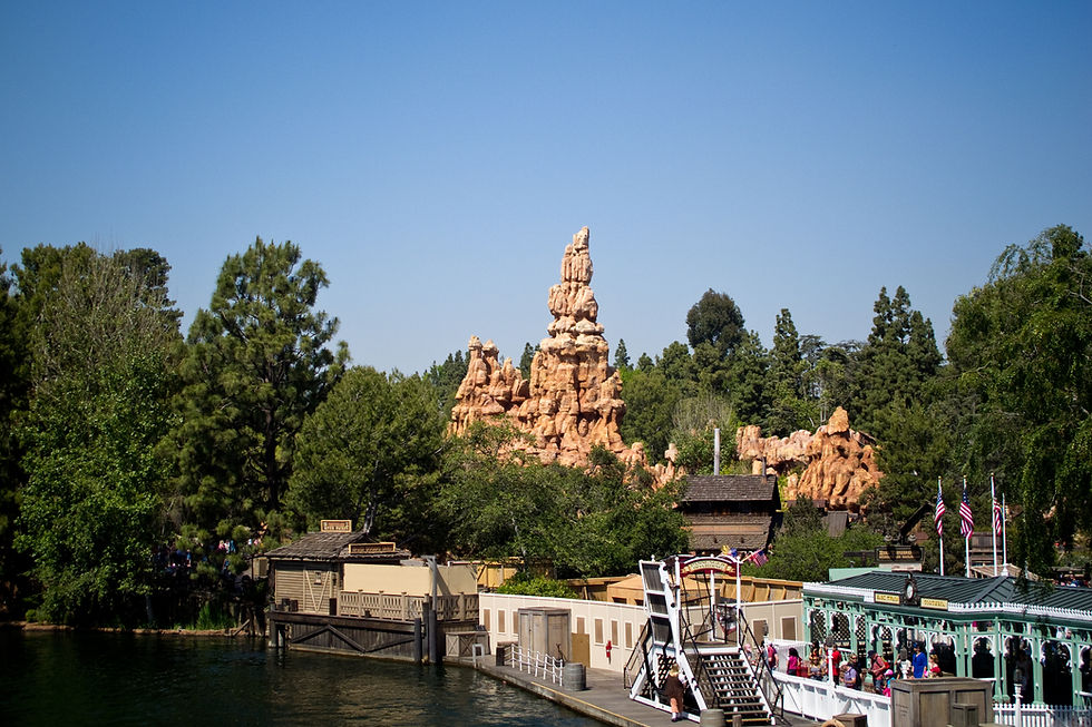 Riverboat docked by a wooden pier, surrounded by trees. Red rock formations in the background. People walking, flags waving under blue sky.
