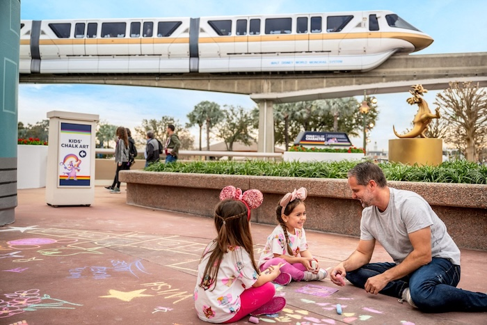 Man and two children draw with chalk on a colorful sidewalk. A monorail passes above. Nearby sign reads "Kids' Chalk Art."
