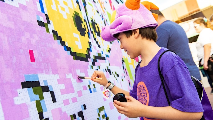 Child in purple shirt and colorful hat paints a large color-by-number mural in vibrant shades. Others in background. Joyful atmosphere.
