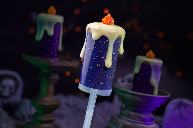 Halloween-themed popsicle designed like a purple candle with dripping white "wax" and orange flame. Spooky bats pattern. Dark background.