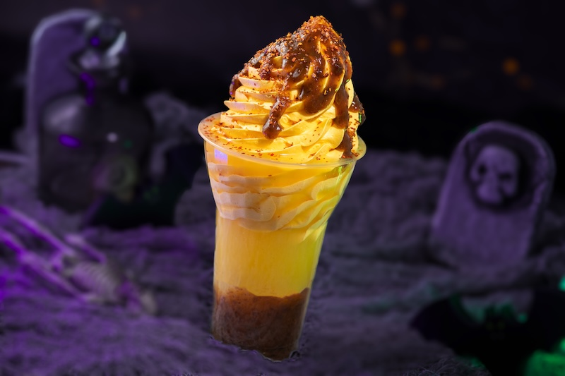 A spooky-themed caramel sundae with whipped topping. Background includes a grave, skeleton, and purple lighting, creating a Halloween vibe.