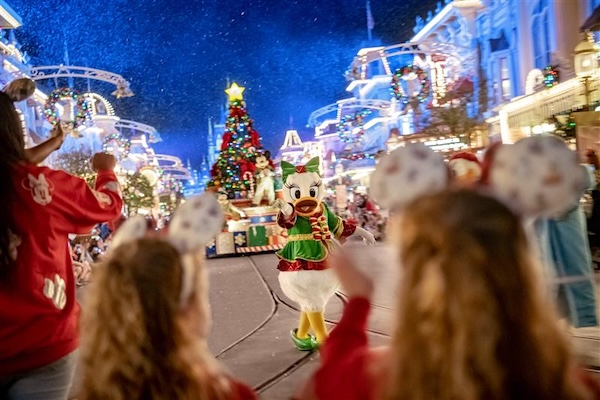 A festive parade with a costumed character waving to children. Decorated street, Christmas tree, and colorful lights enhance the joyful setting.