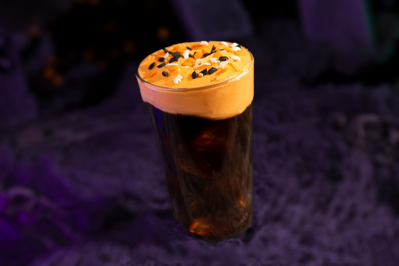 A glass of dark coffee with a creamy orange froth topped with black and white sprinkles, set against a deep purple textured background.