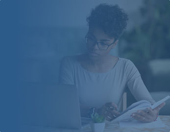 A student wearing glasses studies at a table, looking at a laptop while holding a notebook and pen.