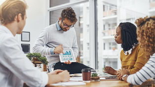 Professional leading a team meeting while presenting business performance data on a tablet in a collaborative office setting