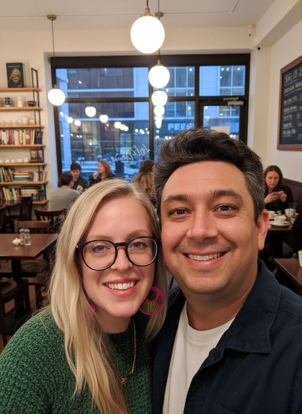 A smiling man and woman taking a selfie inside a café, with bookshelves, hanging lights, and other people sitting at tables visible in the background.