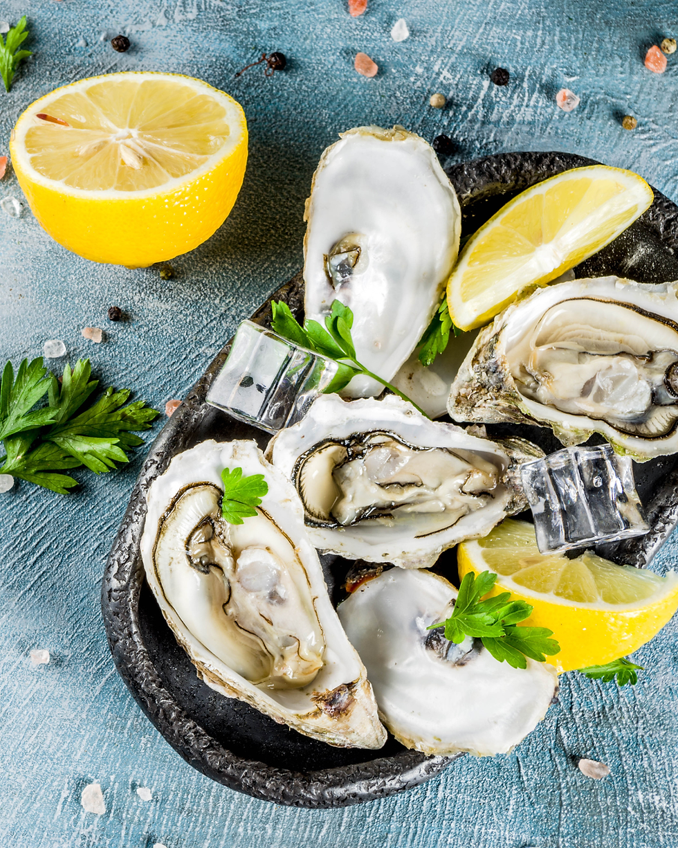 Fresh oysters on a black plate with lemon wedges, parsley, and ice cubes on a textured blue surface. Salt and pepper scattered around.