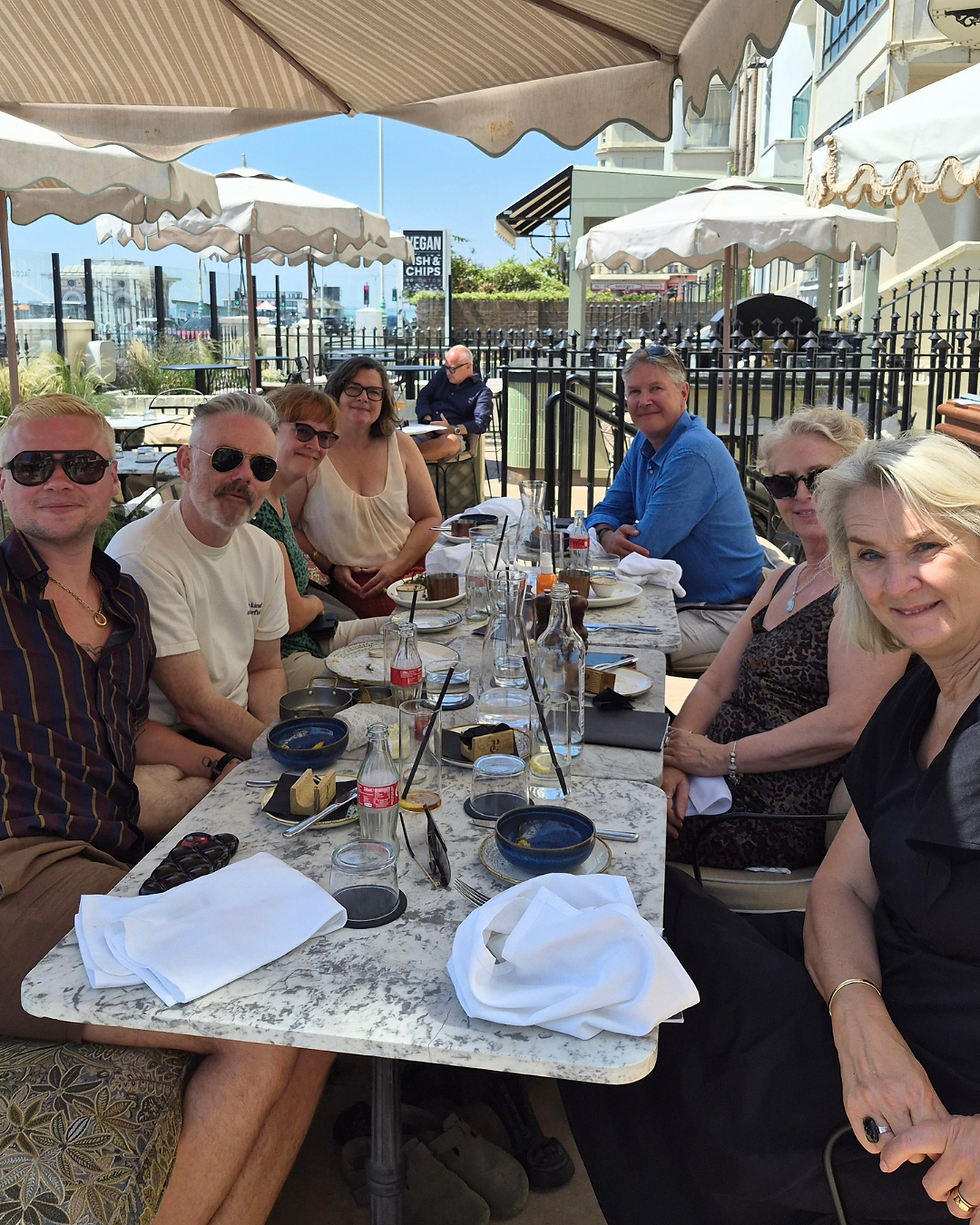 Seven adults smile at an outdoor café table under umbrellas, with empty plates and a VEGAN FISH CHIPS sign in back.