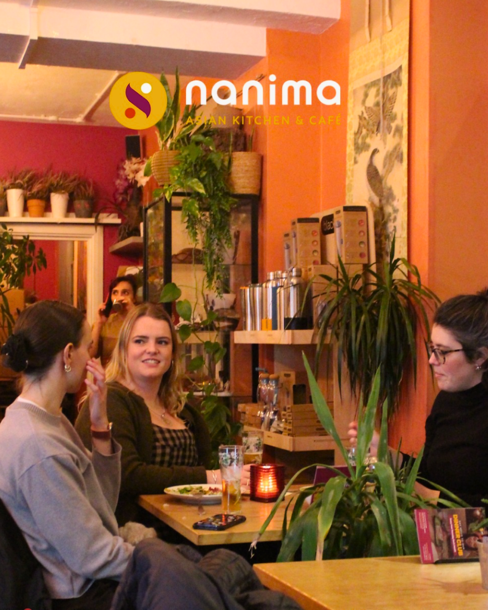 Three women sit at a table in a cozy café with plants. They are conversing, with drinks and a candle on the table. "Nanima" logo visible.