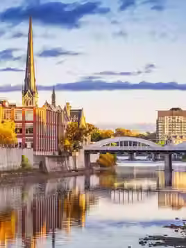 A walkway and bridge along a canal in the city of Cambridge, Ontario, Canada, with autumn leaves and church steeples in the background on either side of the river.