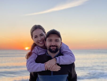 A husband and wife pose for a picture on a beach, with the ocean and a warm sunset in the background.